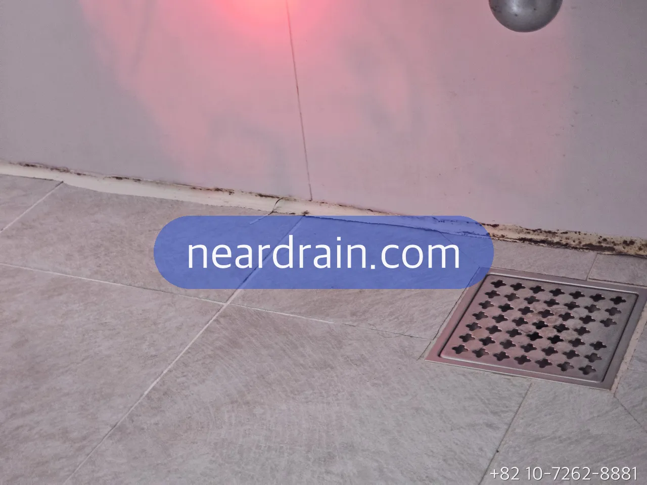 Square floor drain grate in a wet-room bathroom with water pooling around it and dark grout lines at the wall base