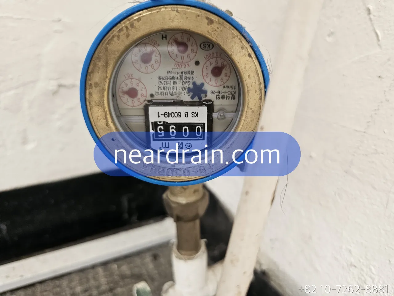 Water meter dial inside a Korean apartment meter box, blue-ringed meter on a vertical supply pipe with the shut-off below it