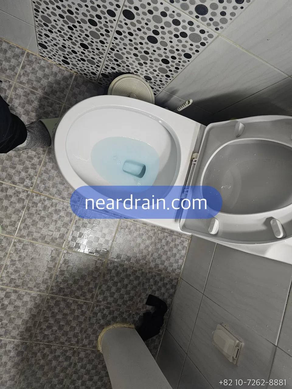 Korean toilet with the bowl water sitting high and blue, tank lid on, next to a washing machine in a small tiled bathroom