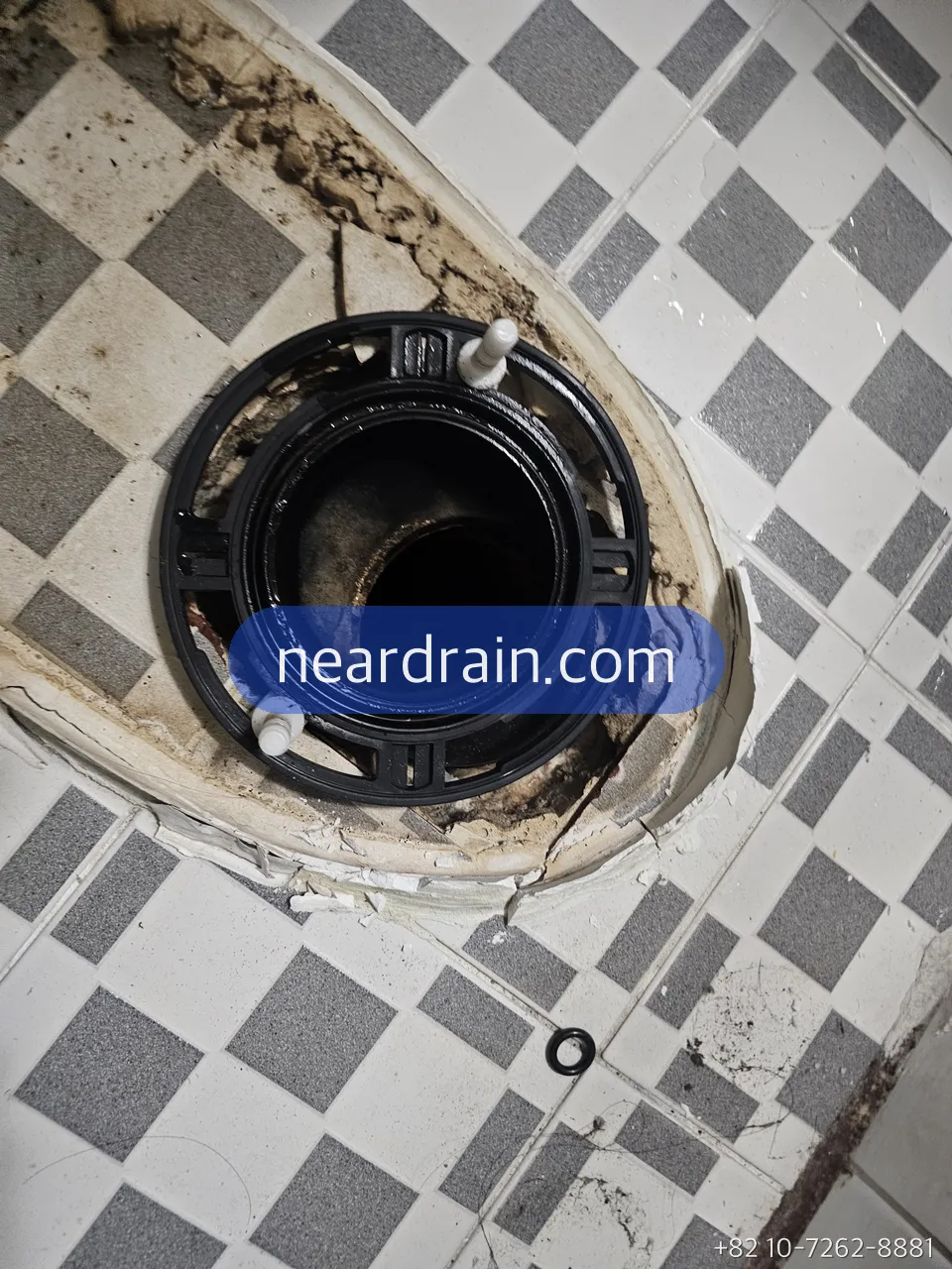 Toilet removed from a chequered-tile Korean bathroom floor, exposing the black floor flange and drain opening